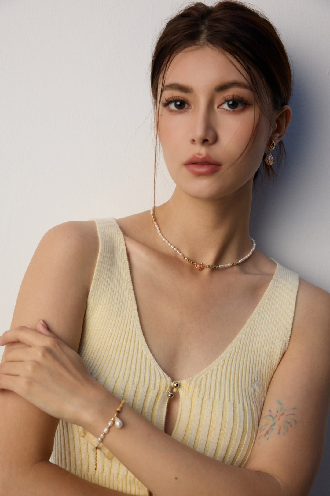 Young woman with straight brown hair gazes confidently. She wears a cream ribbed top, pearl necklace, and bracelet. Subtle floral tattoo on arm.