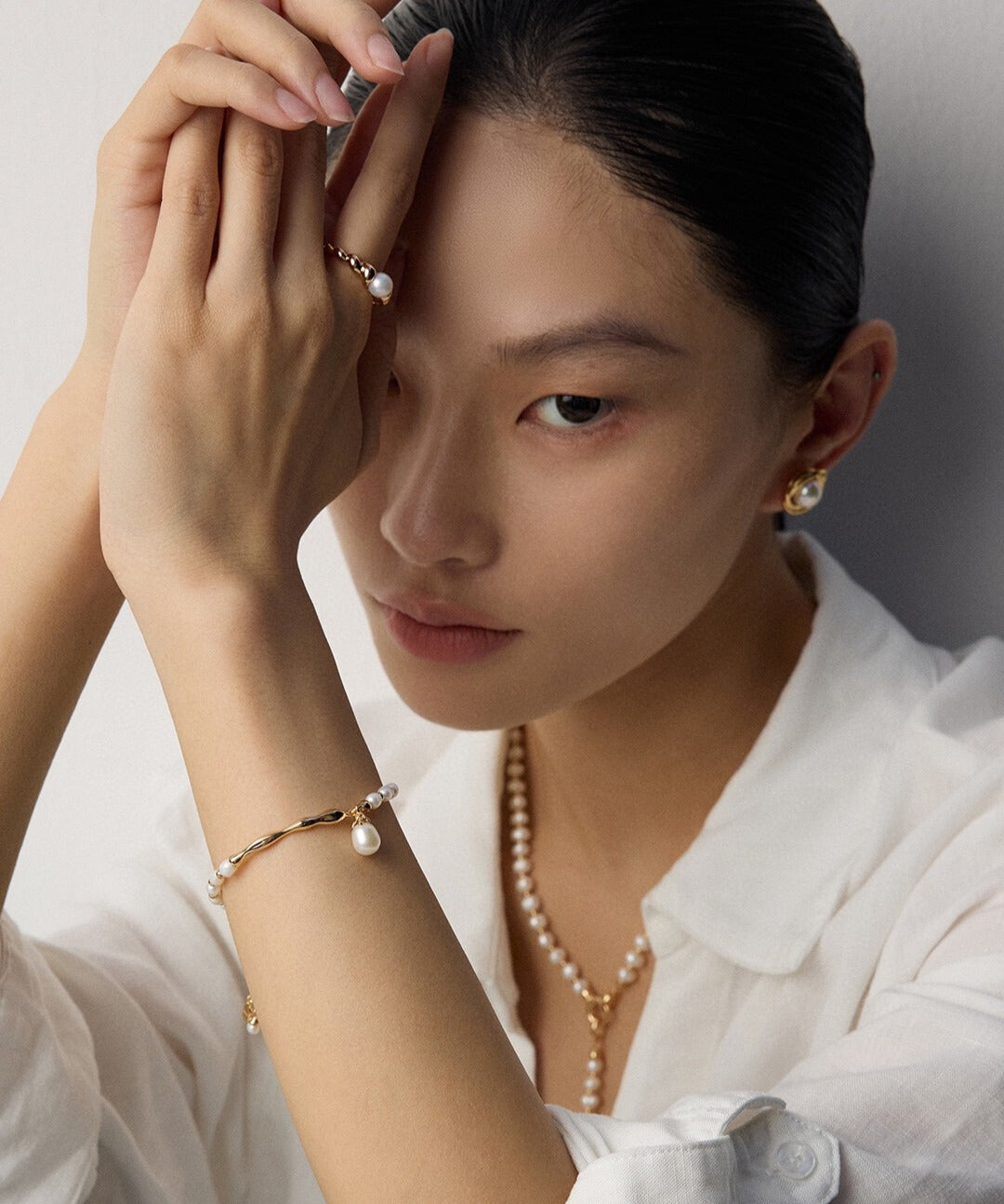A woman in a white blouse poses elegantly, showcasing pearl jewelry, including a bracelet, earrings, and necklace. Her expression is serene and thoughtful.
