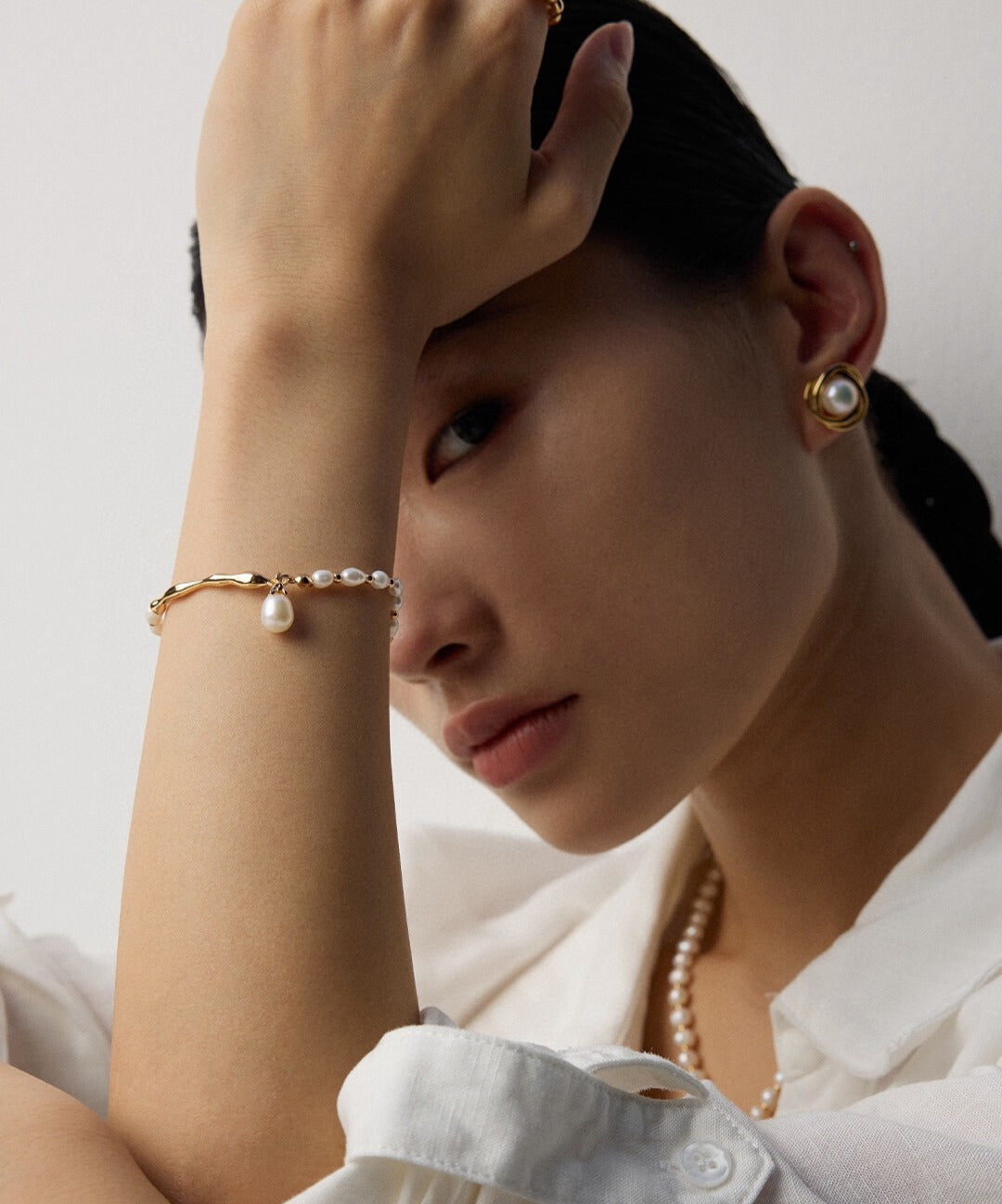A woman leans with her arm raised, wearing pearl earrings and necklace, a gold and pearl bracelet. She gazes pensively, conveying elegance.