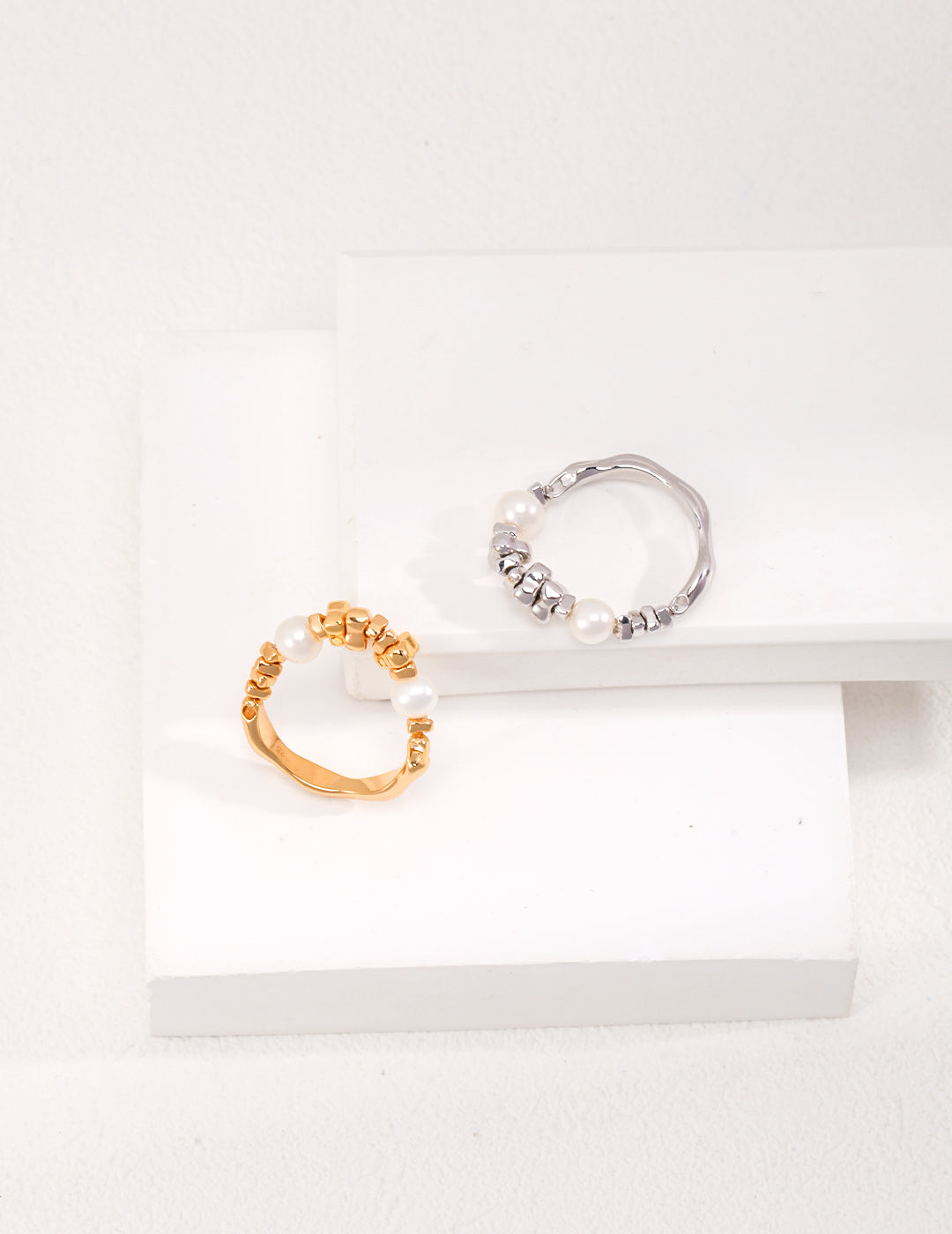 Two rings are displayed on a white surface, showcasing a silver and gold pearls ring.