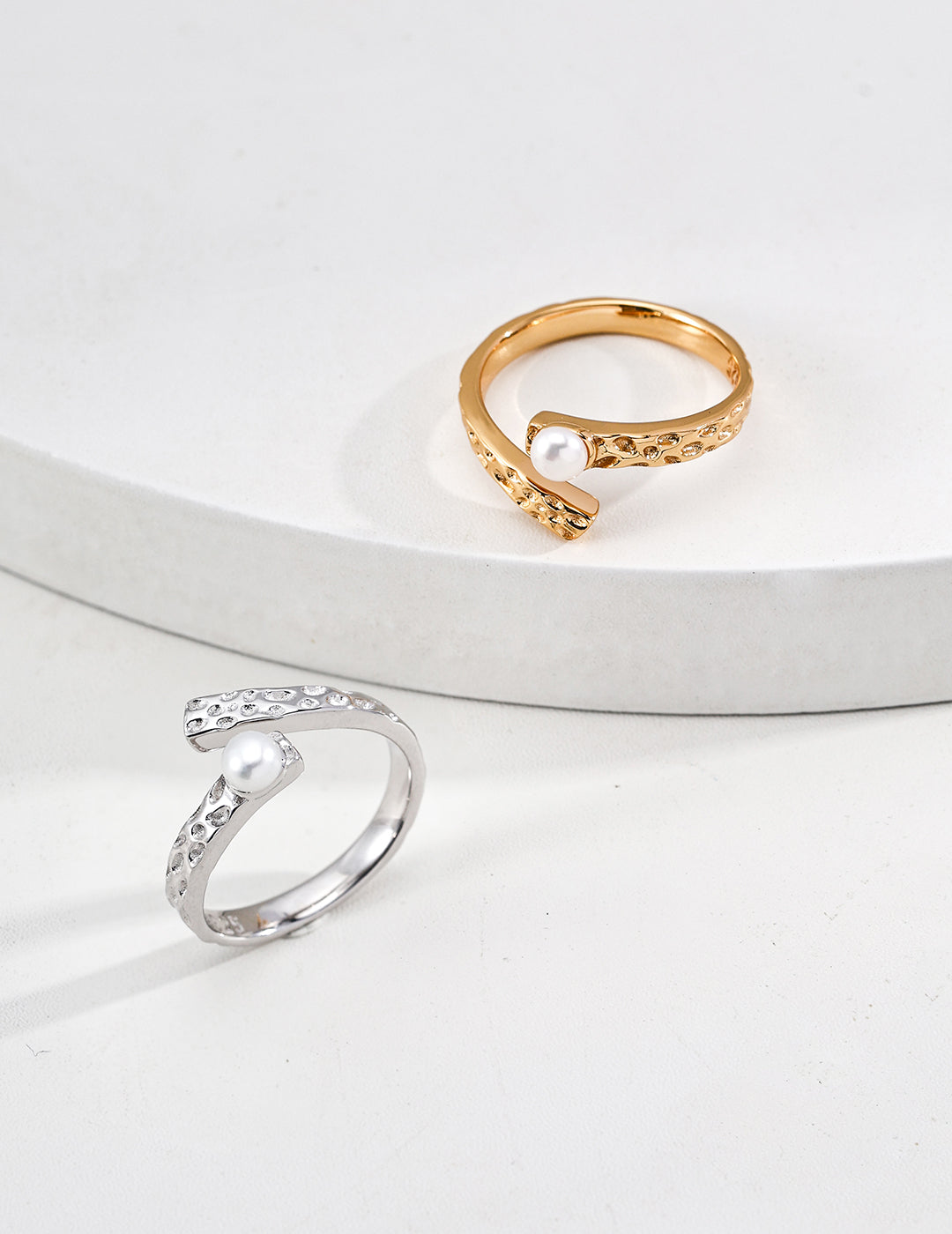 Studio product photograph of the Celine ring displayed in both silver and gold finishes on a minimalist white surface, showcasing the open bypass band with a hammered, dimpled texture and a single white pearl accent.