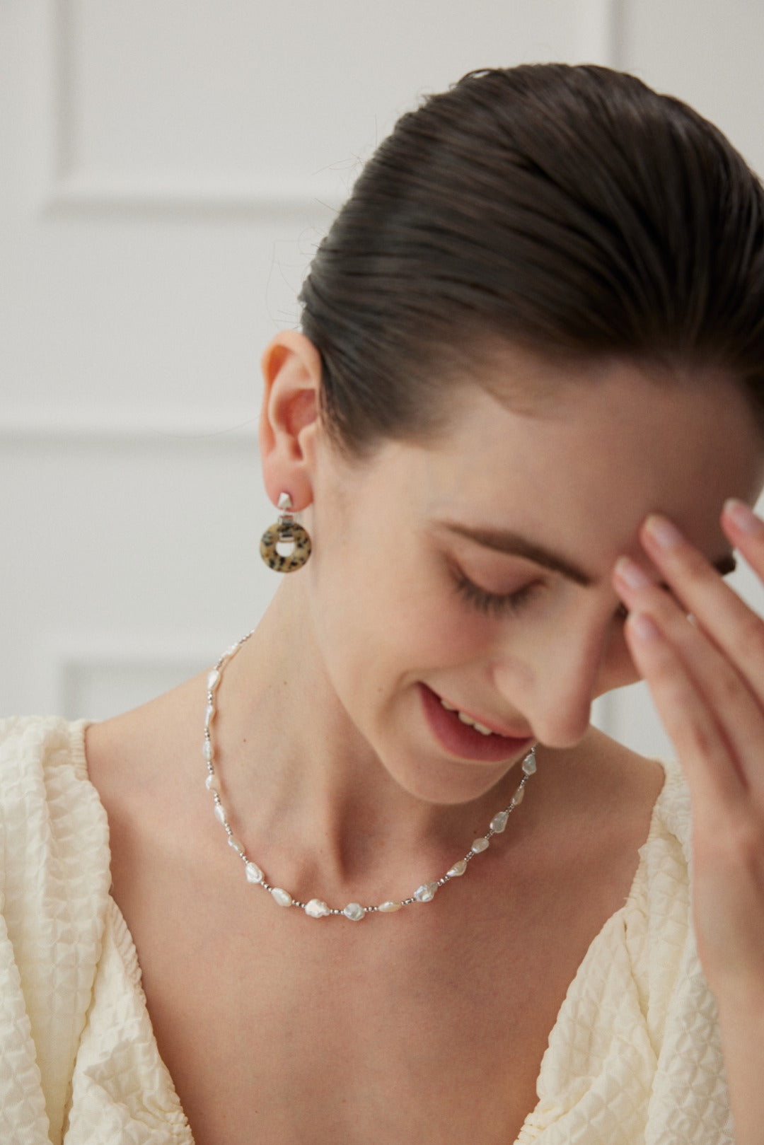 A smiling woman holds her head, showcasing a close-up of her baroque pearl necklace.