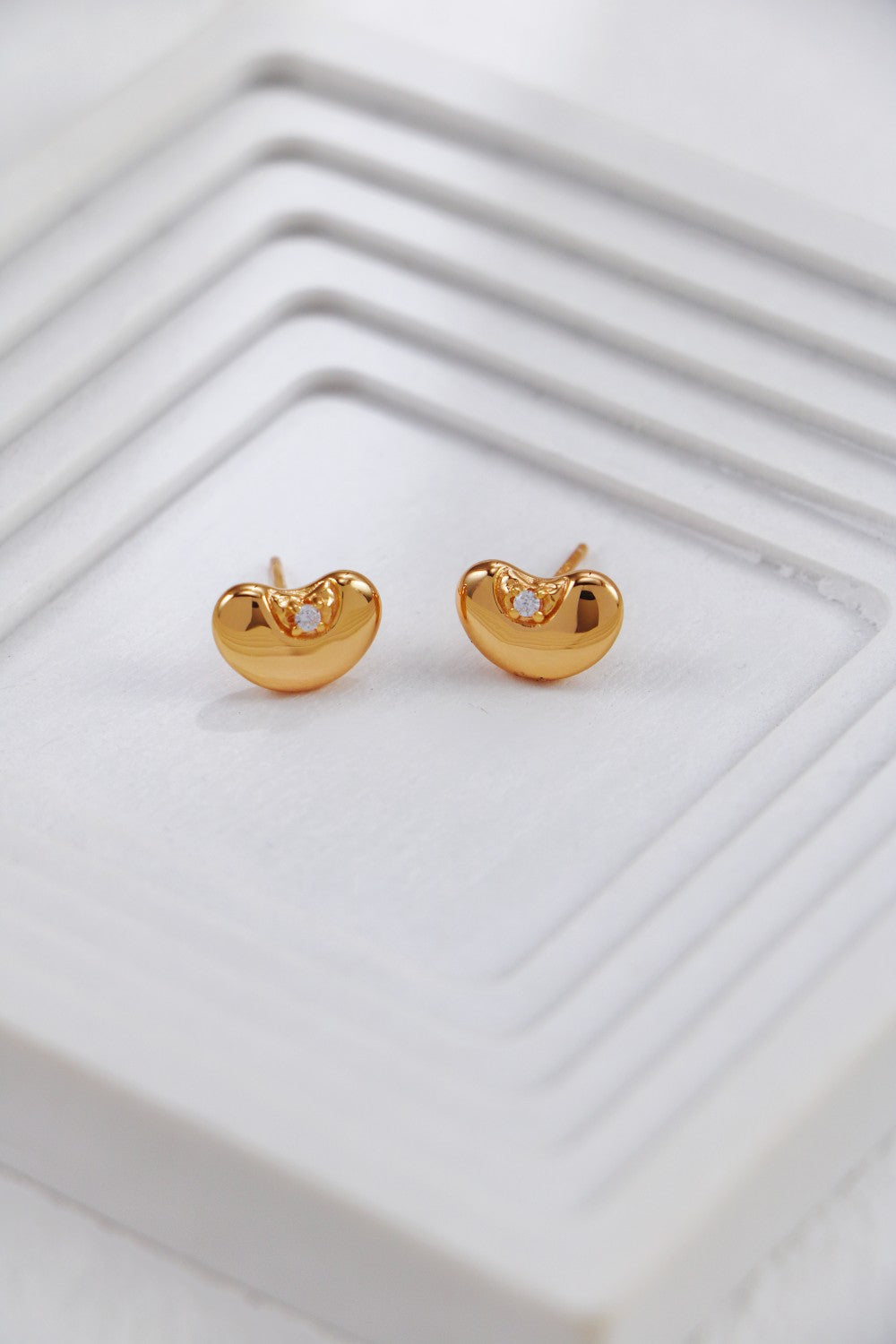 Close-up of a pair of gold-toned stud earrings shaped like rounded beans, each featuring a small inset cubic zirconia at the top center, displayed on a smooth white surface.