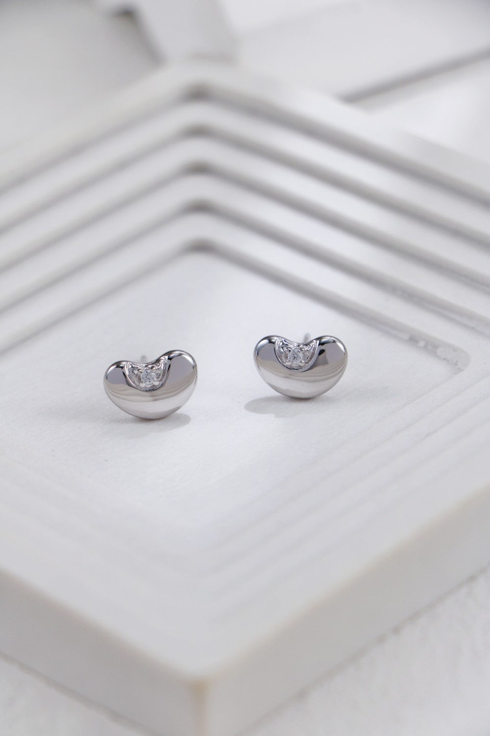 A pair of sleek, bean-shaped silver studs made of S925 silver, each accented with a small, sparkling cubic zirconia, resting on a geometric white textured background.