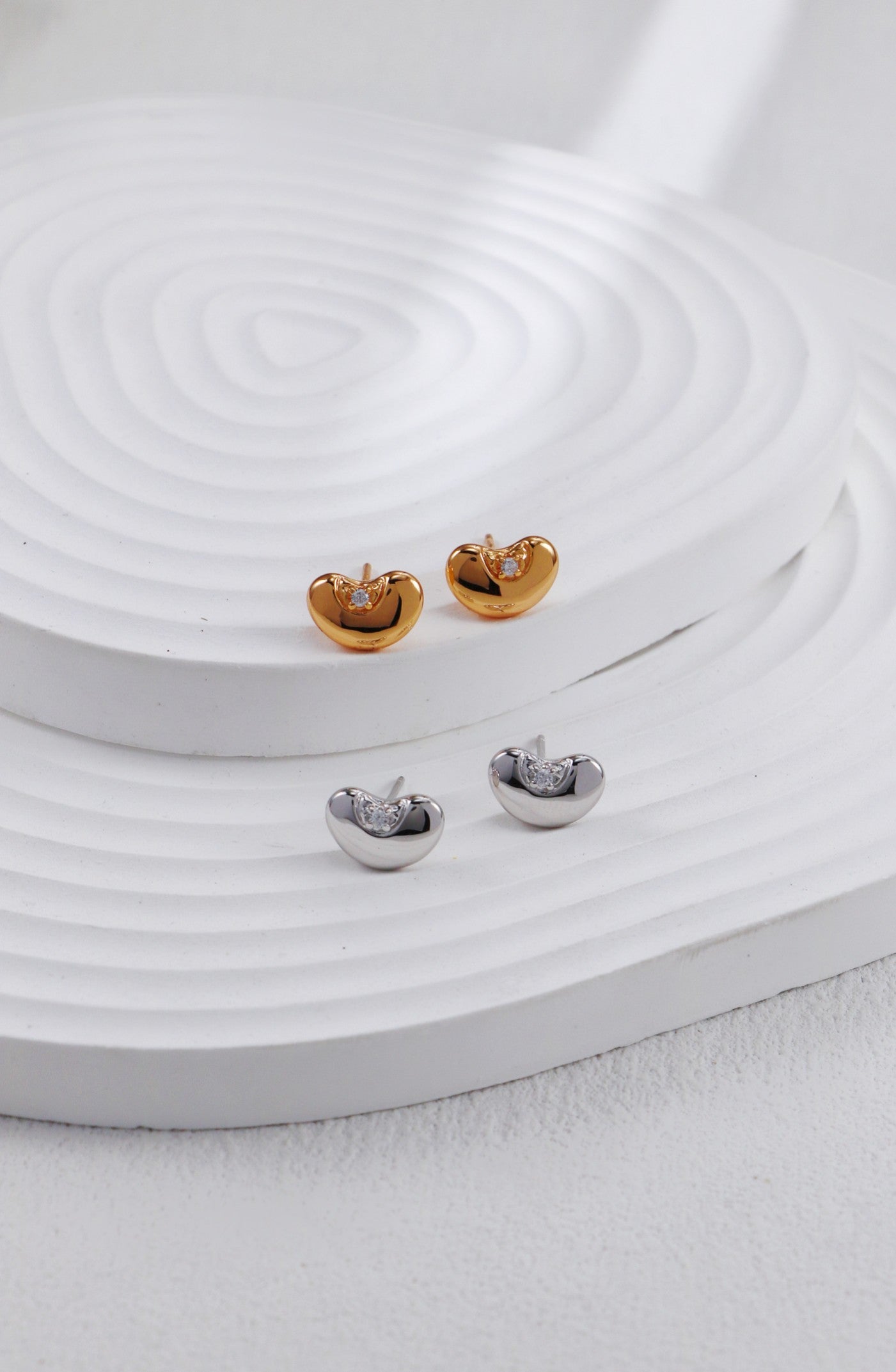 Two pairs of bean-shaped stud earrings—one in warm gold tone and one in polished silver—each set with a single cubic zirconia, arranged side by side on a ridged white platform.