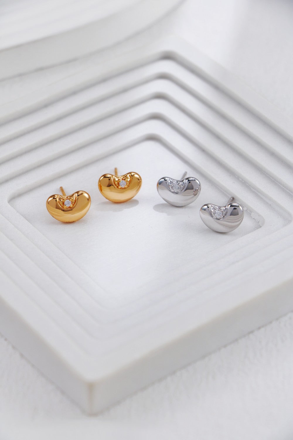Two pairs of bean-shaped stud earrings—one in warm gold tone and one in polished silver—each set with a single cubic zirconia, arranged side by side on a ridged white platform.