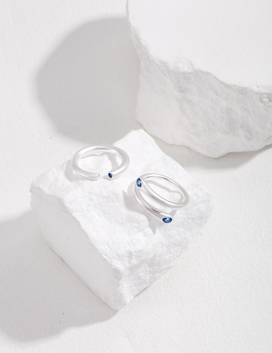 Two uniquely designed silver rings with blue aquamarine stones on a white rock.