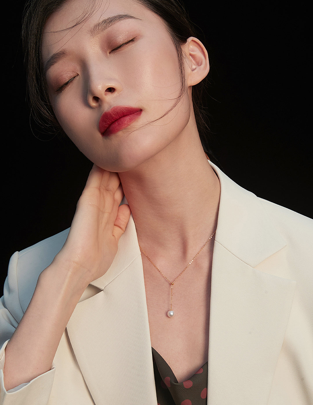 A stylish woman in a white blazer, showcasing red lips and a karat gold necklace with a beautiful freshwater pearl.