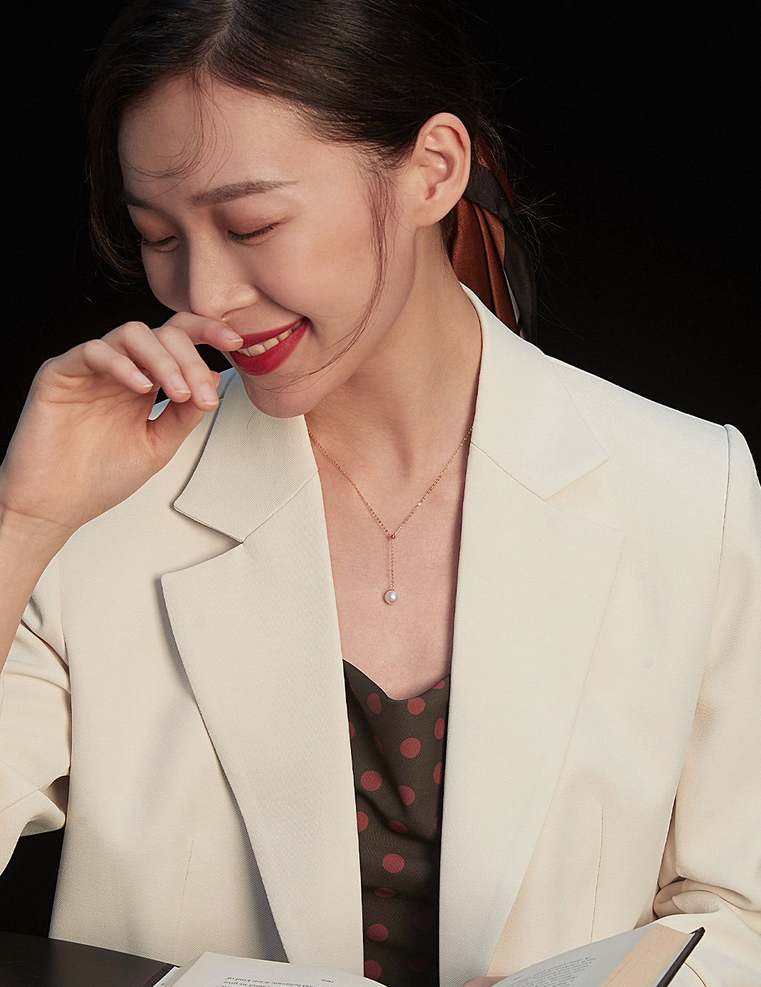 A woman in a white blazer and polka dot shirt sits at a table, engrossed in a book, wearing a gold necklace with a pearl.