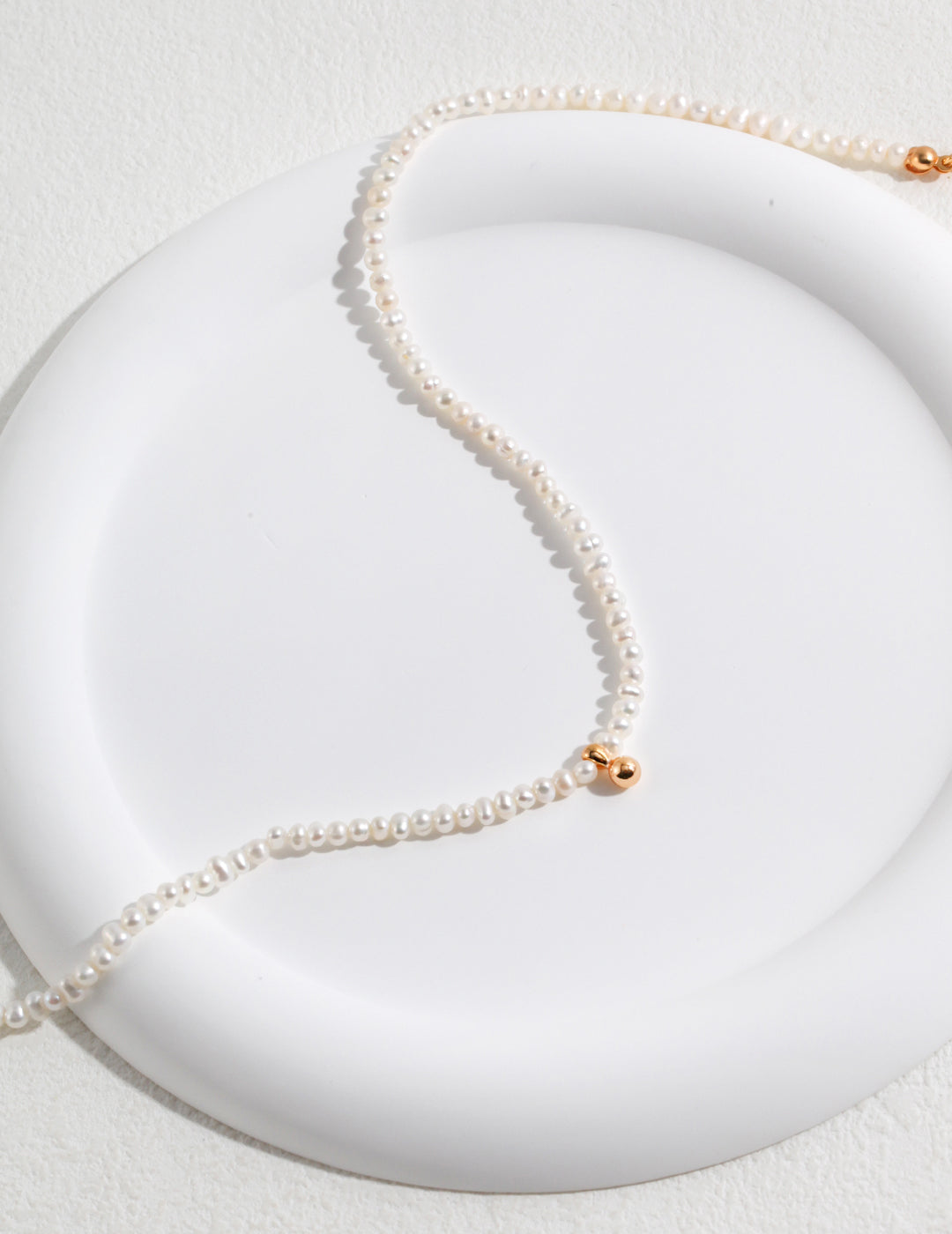A close-up shot of a white plate with a freshwater pearls necklace, featuring a gold plated bead centerpiece.