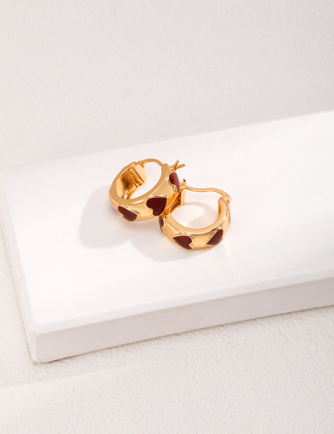 Gold plated hoop earrings with red enamel hearts, adding a touch of elegance and romance to any outfit.