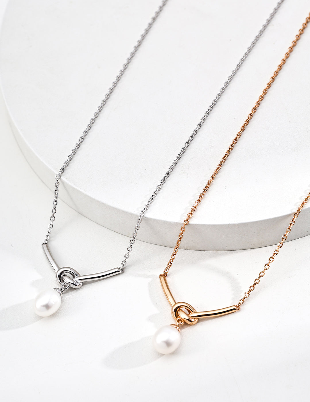 Studio close-up flat lay of the Love Me Knot necklaces side-by-side on white geometric steps, showcasing the curved knot setting and teardrop pearl pendant in both silver and gold finishes.