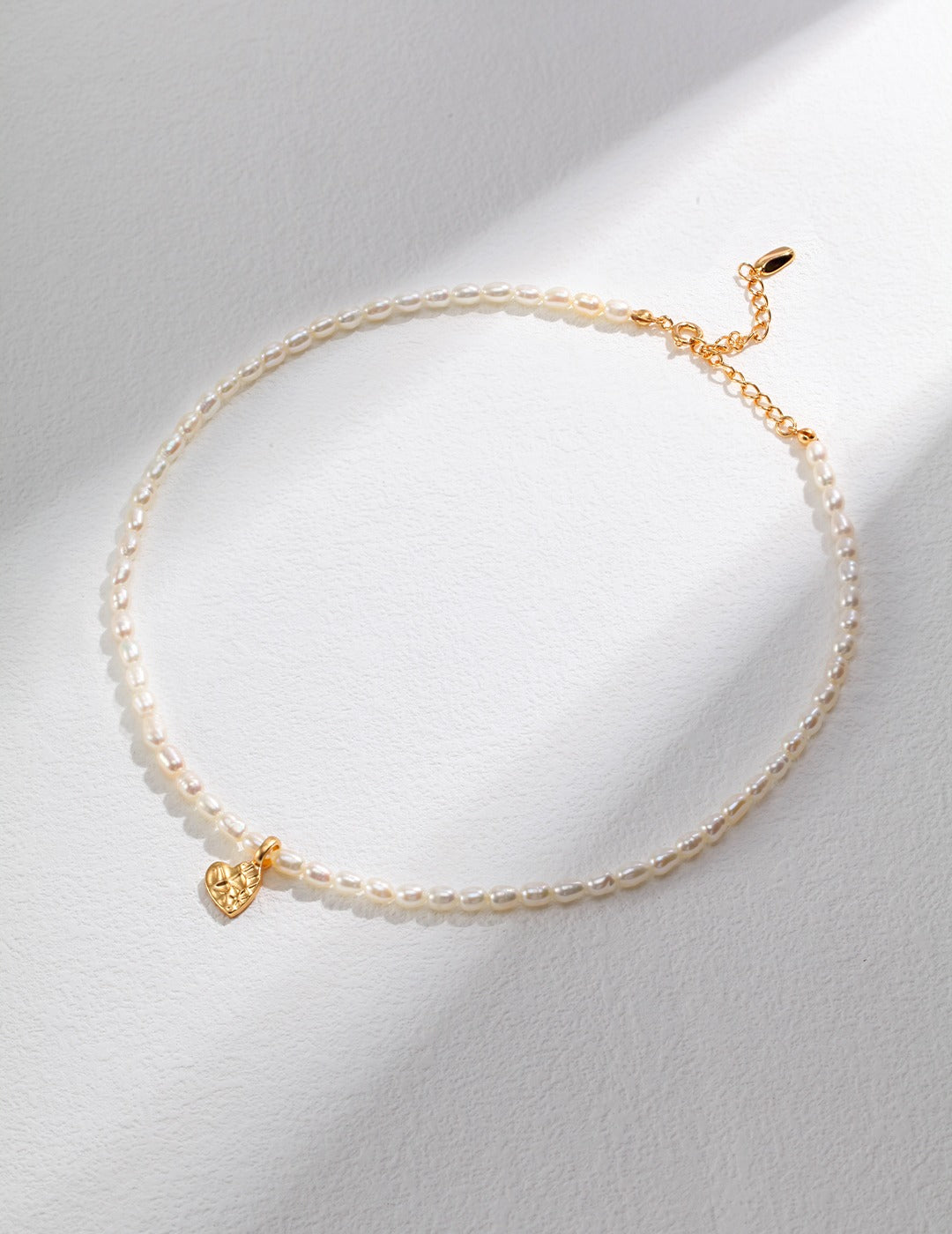 A delicate gold and white pearl necklace with a small heart pendant, displayed on a white surface.