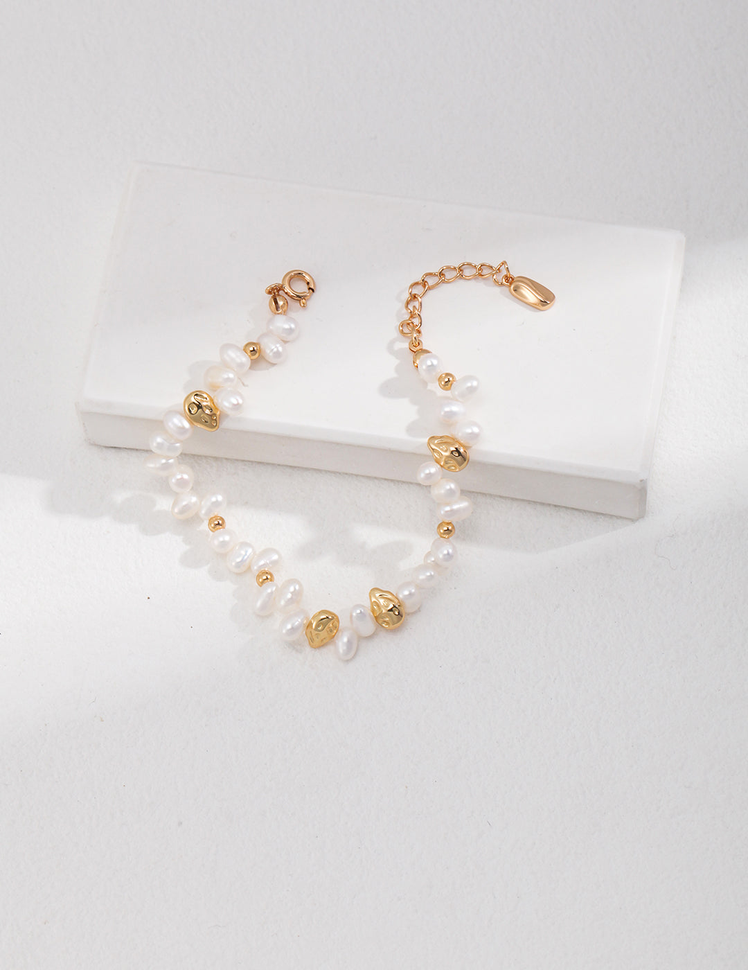 Studio flat lay view of the Natasha pearl and gold nugget bracelet arranged in an open curve across a clean white rectangular display plinth.