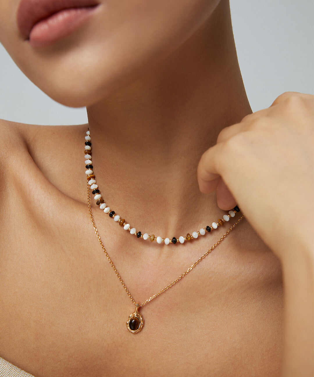 A woman wearing a gold pendant necklace, layered with other necklaces, featuring a tiger's eye gemstone.