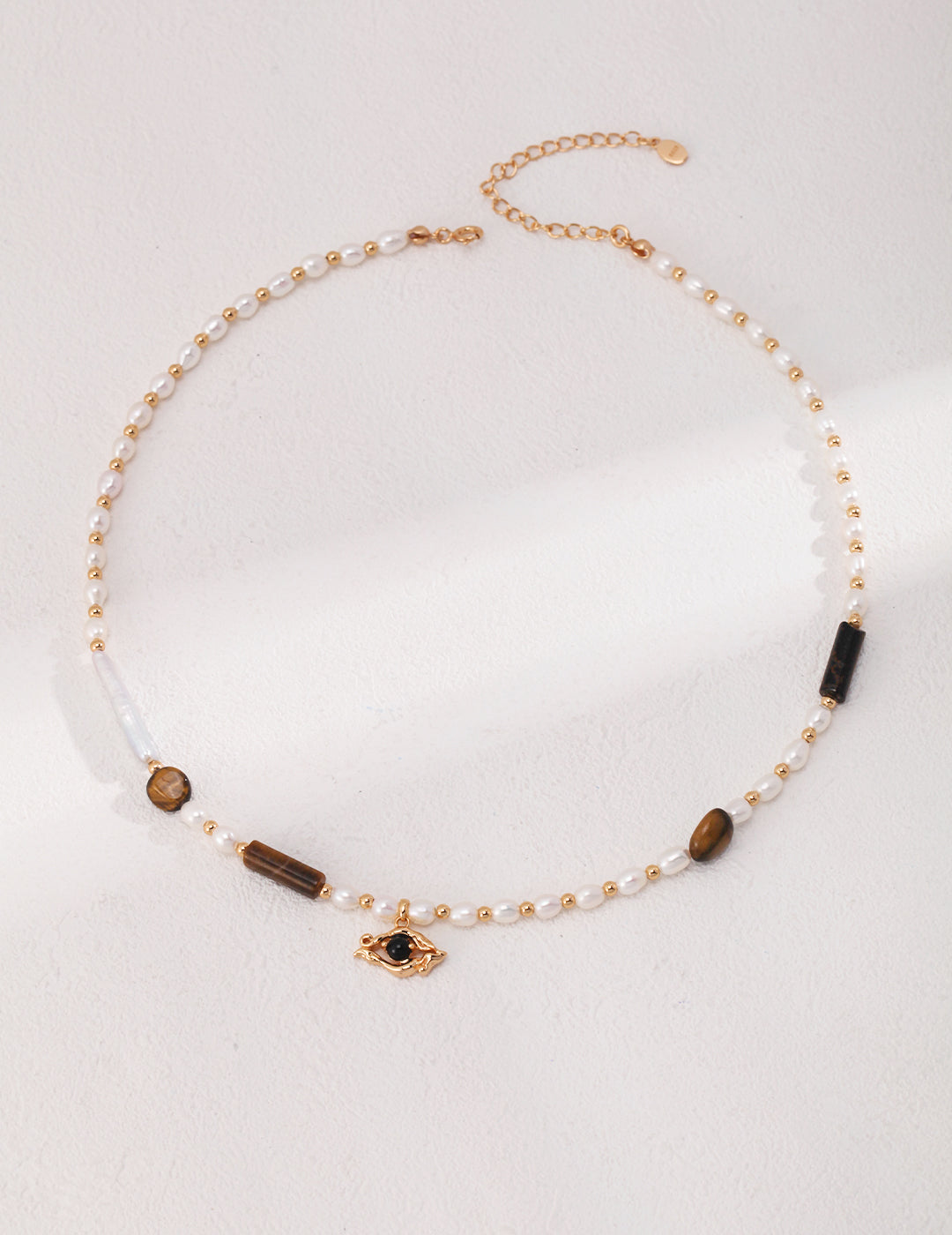 A captivating evil eye necklace adorned with gold beads and pearls, featuring tiger's eye stones, freshwater pearls, and a black agate pendant.