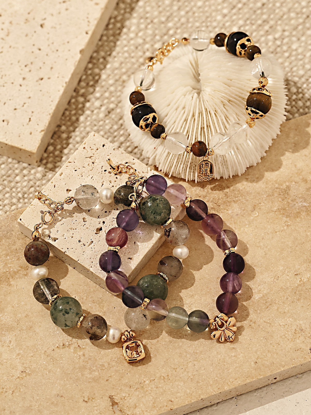 Two beaded bracelets featuring gold charms on a textured surface, alongside a delicate sea shell.