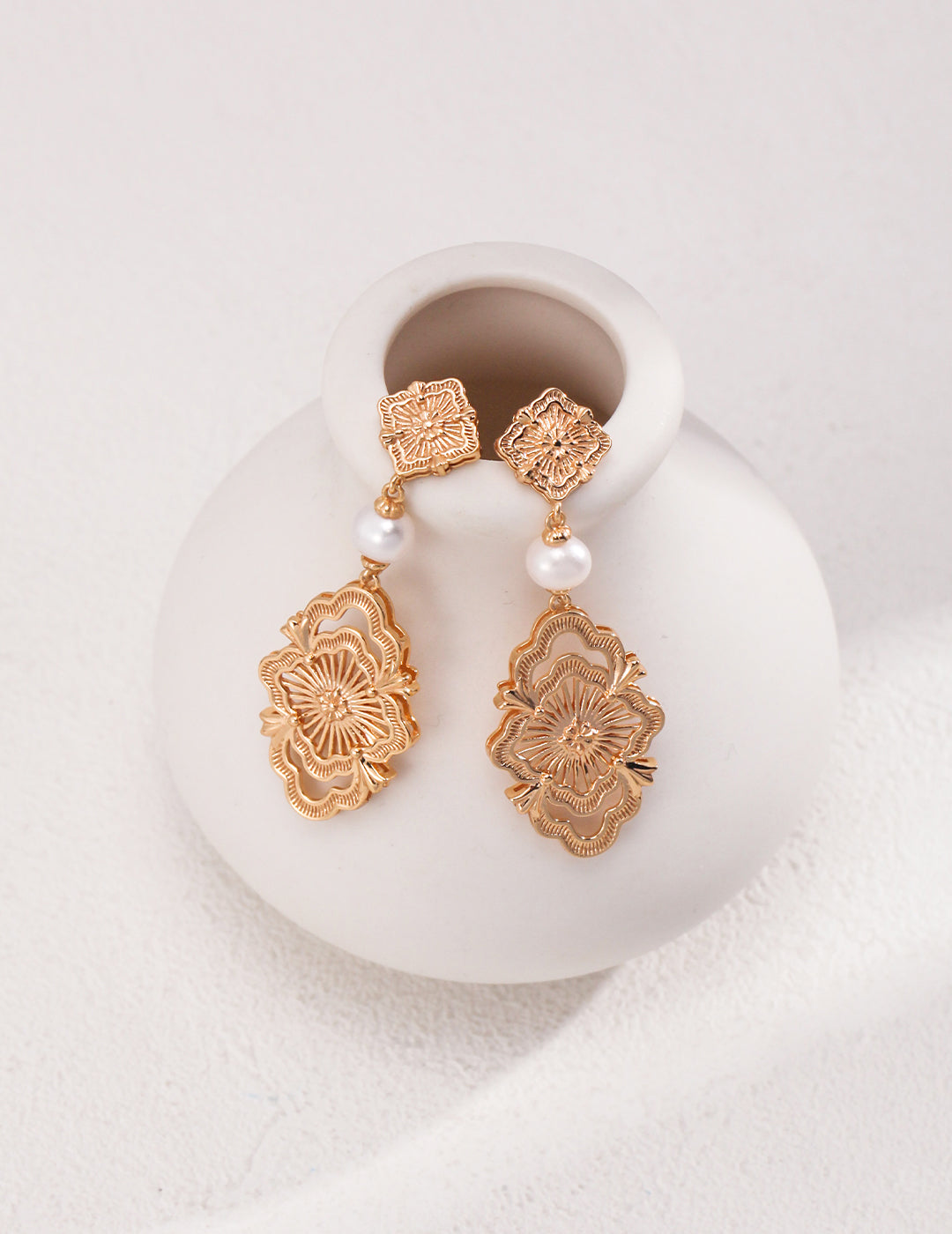 Gold earrings with freshwater pearls elegantly placed on a white vase. A touch of nobility and beauty.