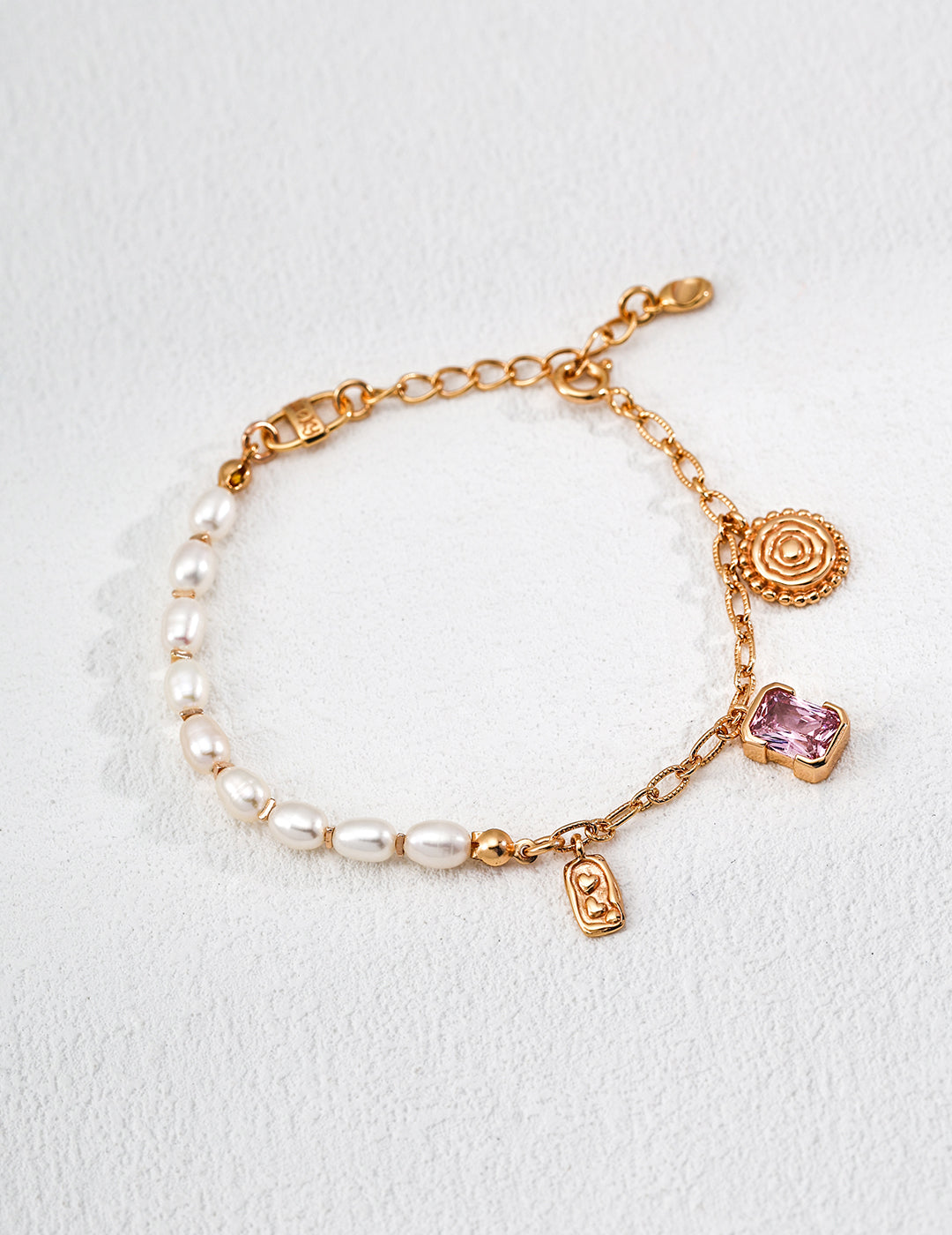 Gold chain Rebecca bracelet with freshwater pearls, a round textured gold charm, a small rectangular gold charm, and a pink gemstone girly charm, displayed on a light textured surface.