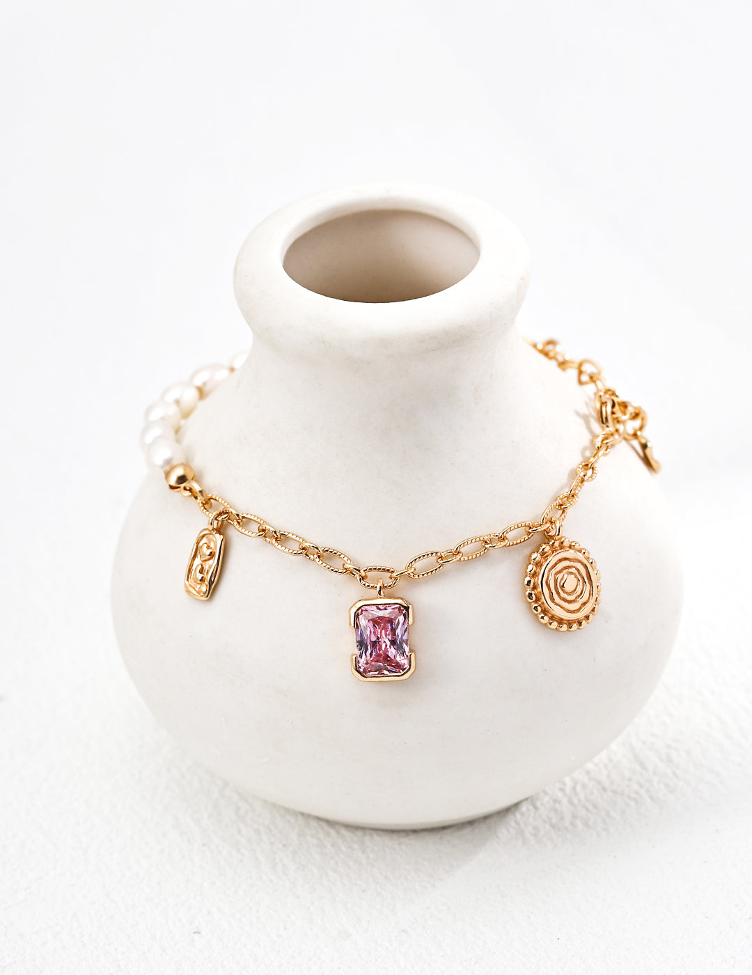 A delicate gold bracelet with a pink zirconia stone, two charms, and pearls drapes over a small, white ceramic vase. The setting is minimalist and elegant.