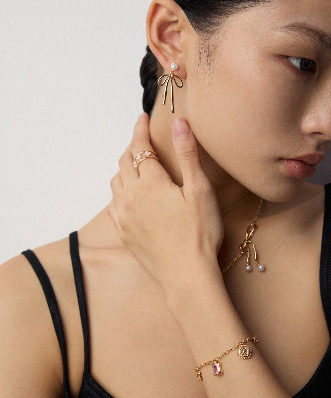 A woman wearing pearl and gold bow earrings, a delicate necklace, a dainty pearl bracelet with charms, and a ring. She looks serene, with a stylish black top.