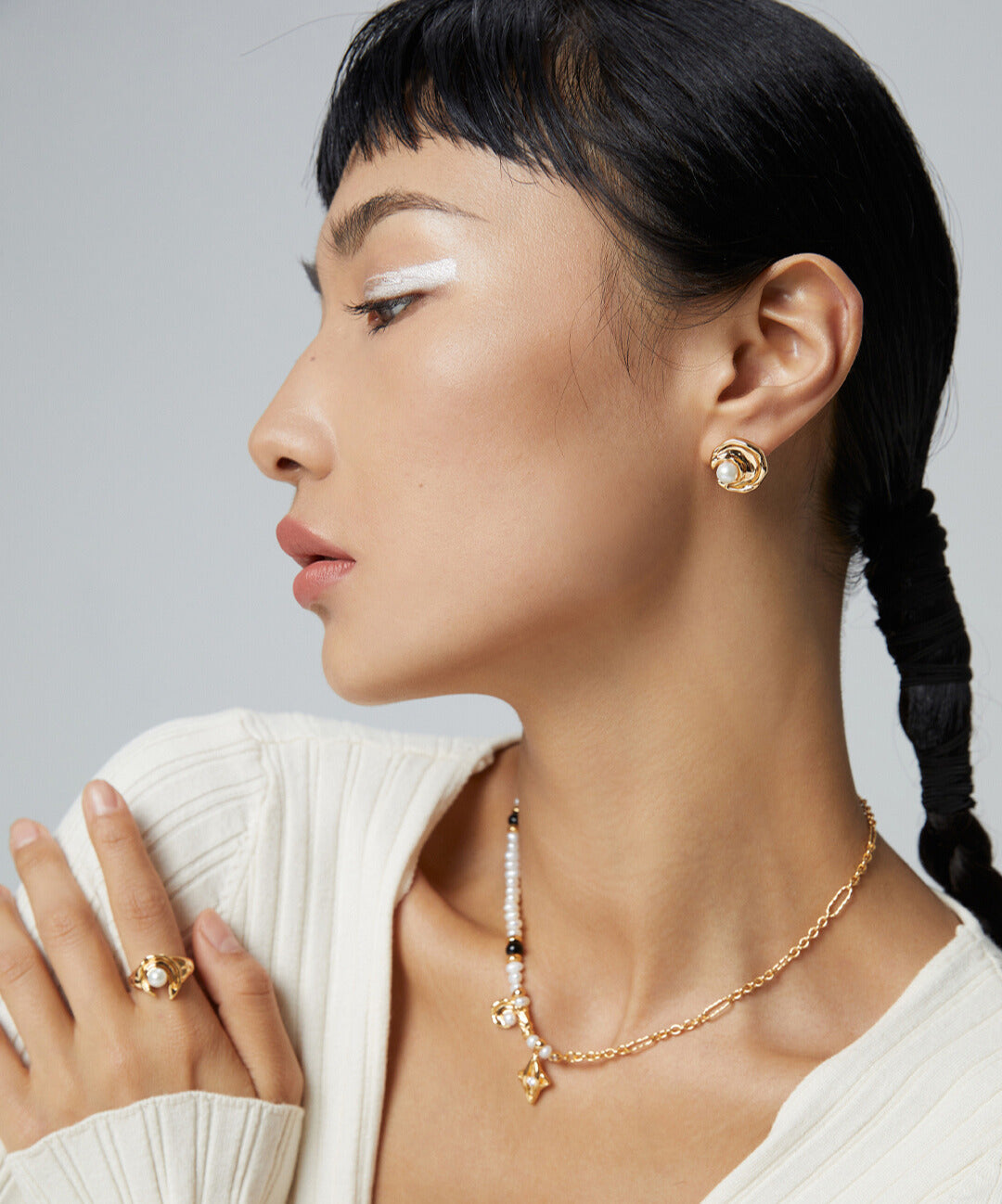 A stylish woman in a white sweater, adorned with elegant gold jewelry including a celestial-inspired necklace with freshwater pearls.