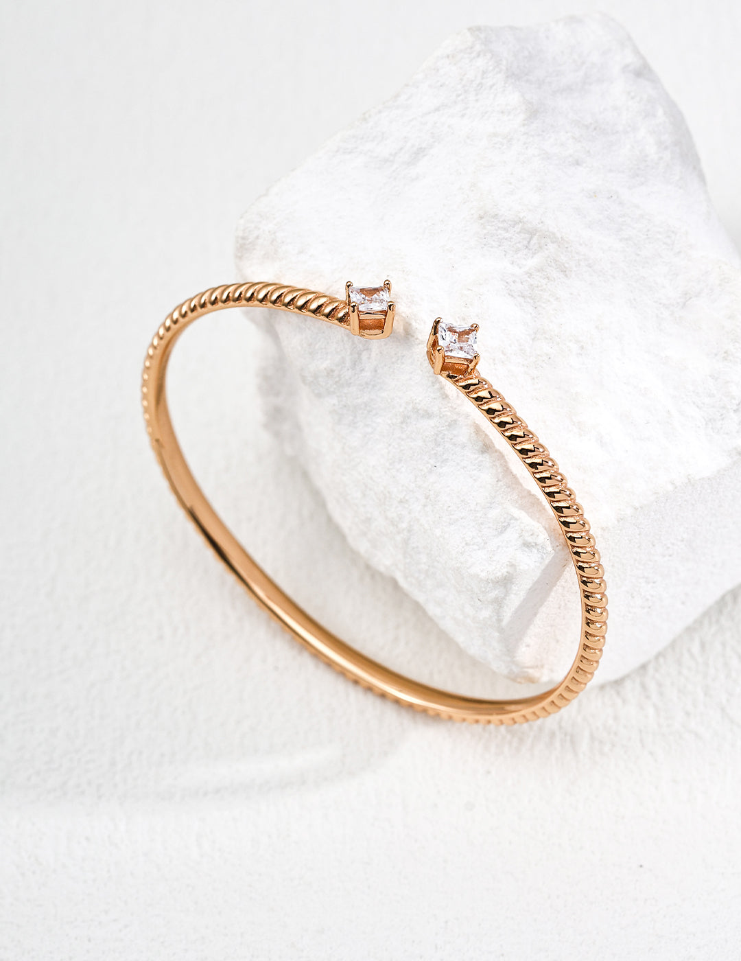 A gold bangle with a textured band and two square-cut zirconia, elegantly wrapped around a rough white stone, set against a soft, neutral background.