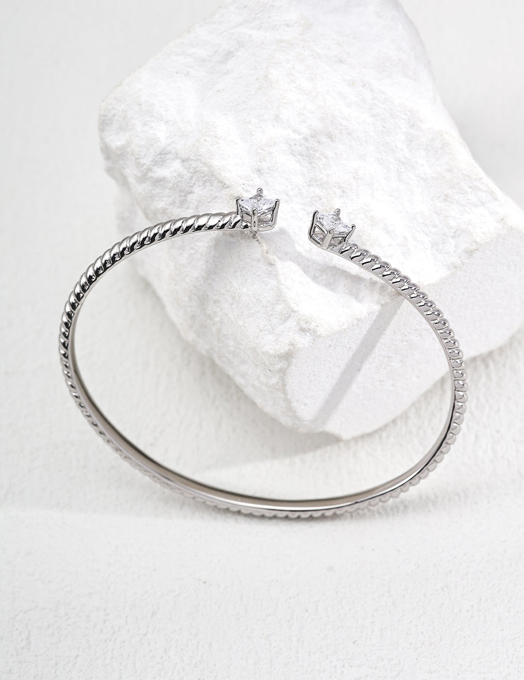 A sleek silver bangle with a twisted design rests elegantly on a textured white stone. The bangle's ends feature small, shimmering gems, conveying elegance and simplicity.
