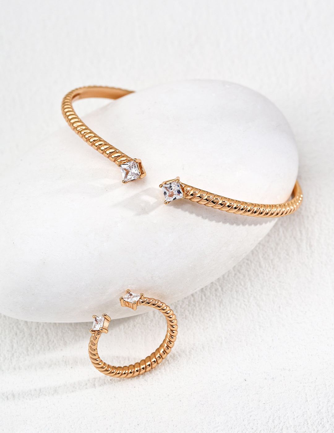 Gold jewelry set with square-cut clear zirconia stones displayed on a smooth white stone. The set features an elegant bangle and matching ring.