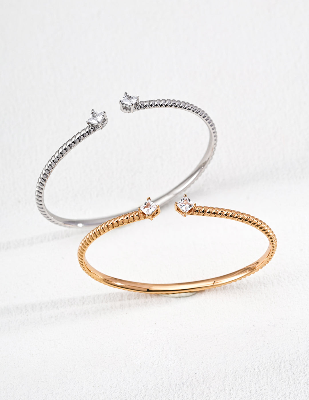 Two elegant bangle bracelets with square-cut gemstones, one in silver and the other in gold, are displayed on a white textured background, conveying luxury and simplicity.