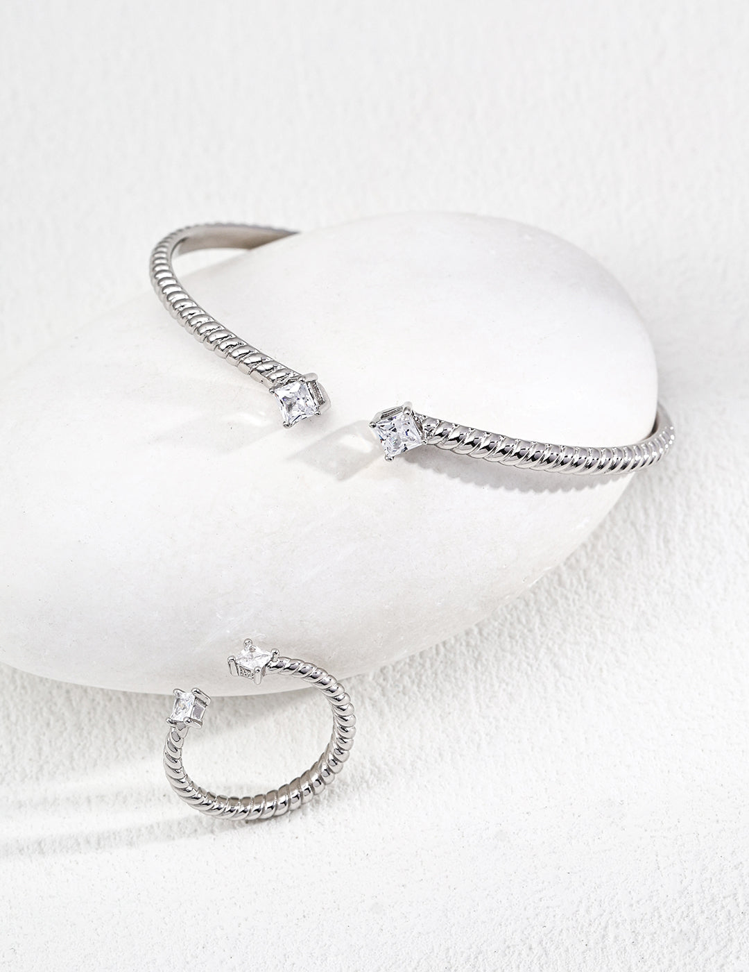 Silver jewelry set with geometric designs, including a bangle bracelet and a band ring, featured on a smooth white stone against a soft, textured backdrop.