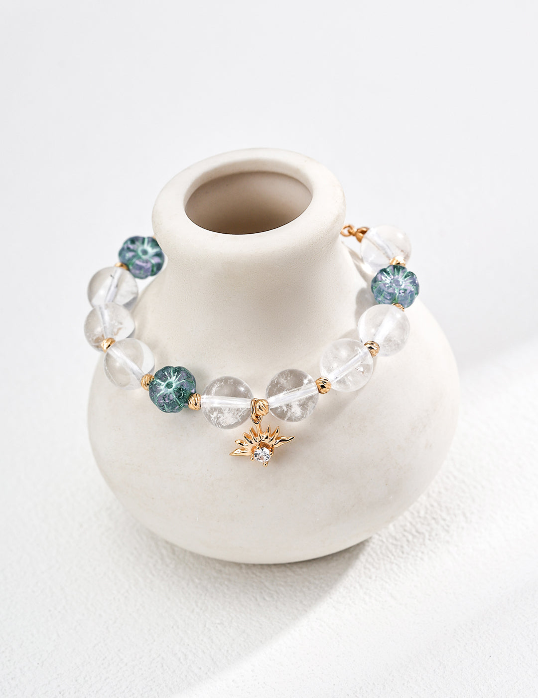 A white vase holds a bracelet featuring blue and green stones, including clear quartz and flower-shaped glass beads.