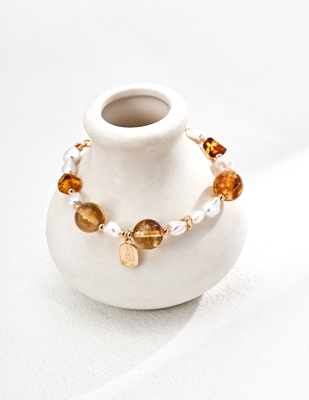 A white vase holds a bracelet made of gold, amber, tiger's eye, and pearls, showcasing a blend of elegant colors.