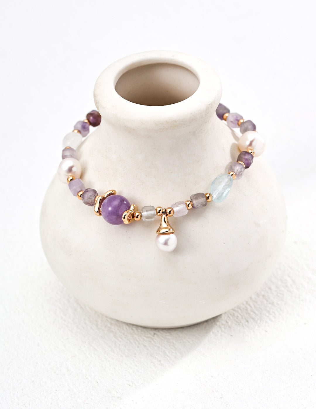 A white vase holds a bracelet made of amethyst and icy aquamarine beads, showcasing purple and white colors.