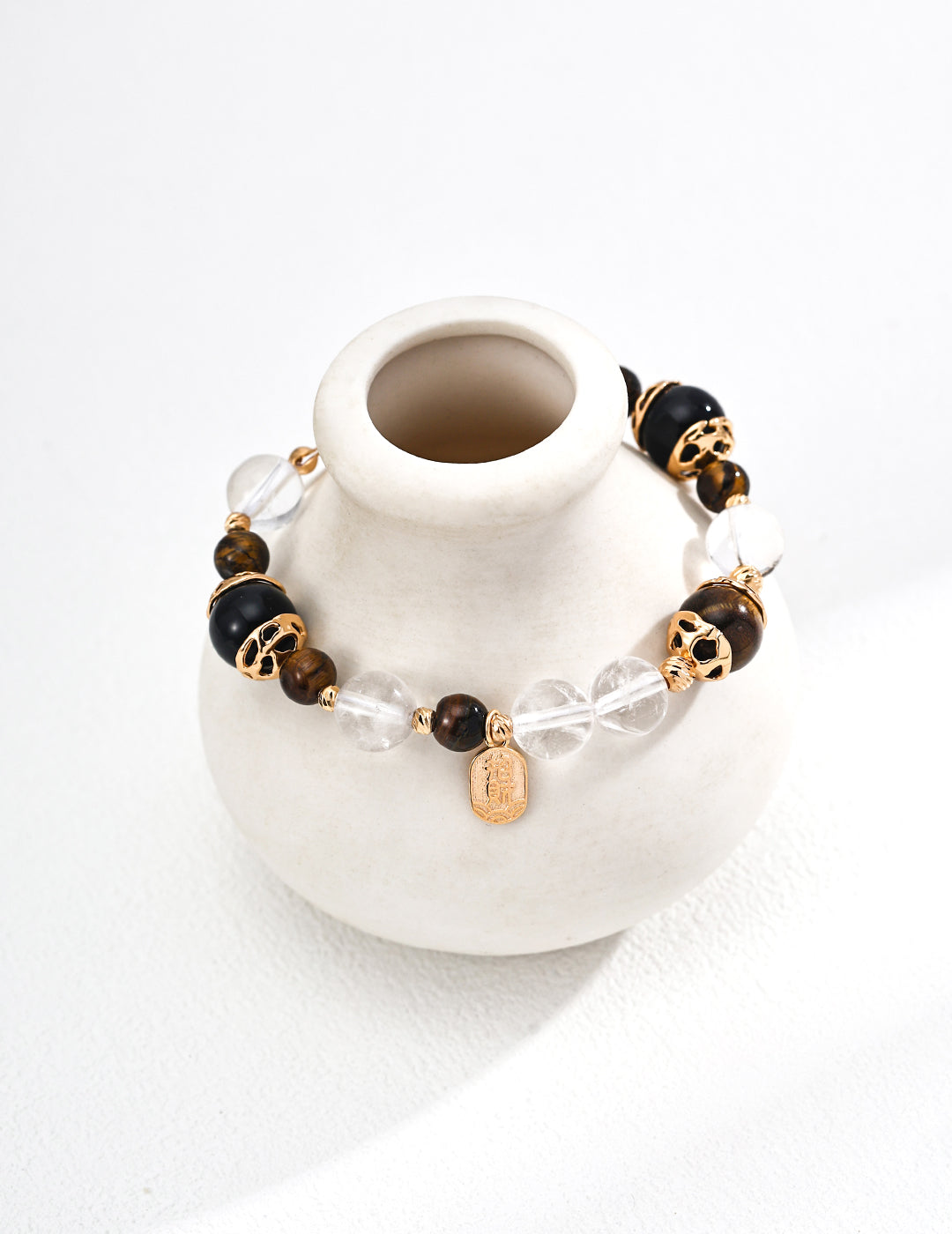 A stylish bracelet made of black and brown beads, clear crystals, gold accents, and a small pendant, resting on a white ceramic vase.