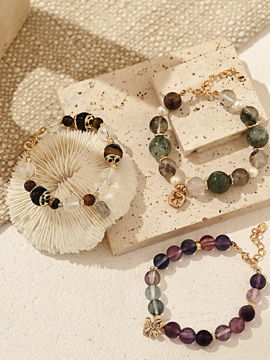 Three elegant beaded bracelets featuring various gemstones and gold accents on textured surfaces, showcasing unique colors and designs.