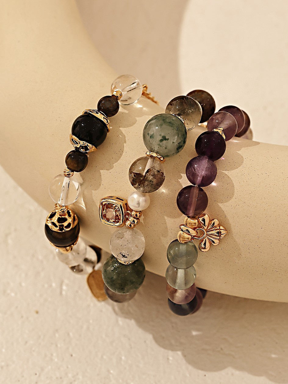 Three elegant bracelets made of various colorful gemstones and pearls, displayed on a neutral-toned curved surface.