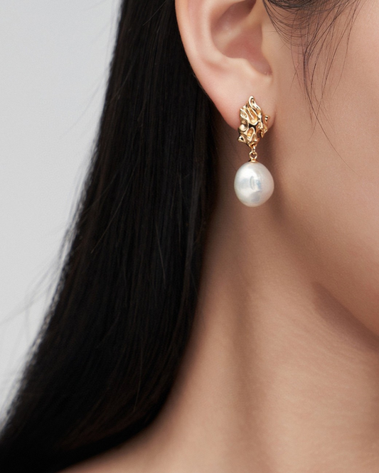 A woman showcases beautiful baroque pearl and gold earrings in a close-up, highlighting their expensive and chic design.

