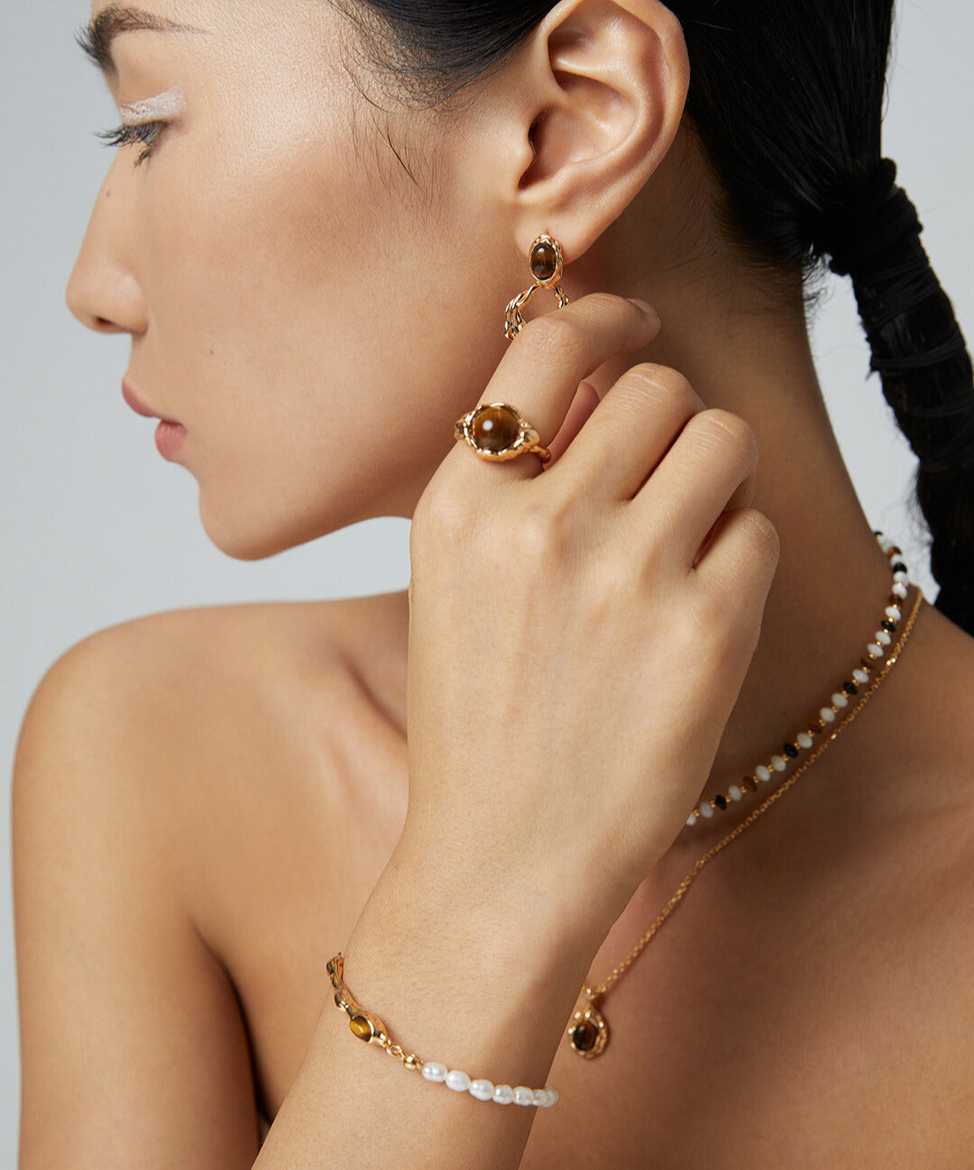 A model wearing a gold necklace and earrings, with a gold plated bangle adorned with freshwater pearls and a tiger's eye centerpiece.