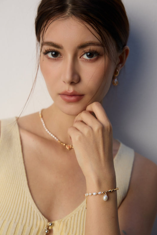 Young woman with thoughtful expression, wearing pearl jewelry, a beige sleeveless top, and subtle makeup. Soft lighting enhances her serene demeanor.