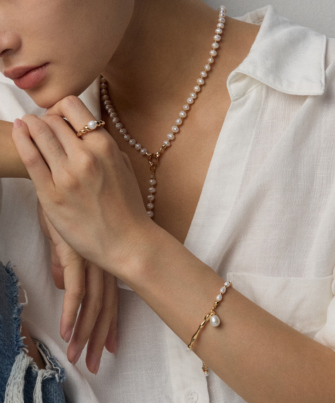 A woman wears a pearl necklace, bracelet, and ring, paired with a white shirt. Her hand is elegantly posed, conveying a sense of sophistication and style.