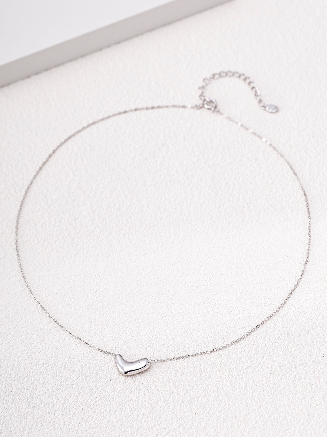 Flat lay of the silver Mezuille Beanie Necklace on a neutral textured background.
