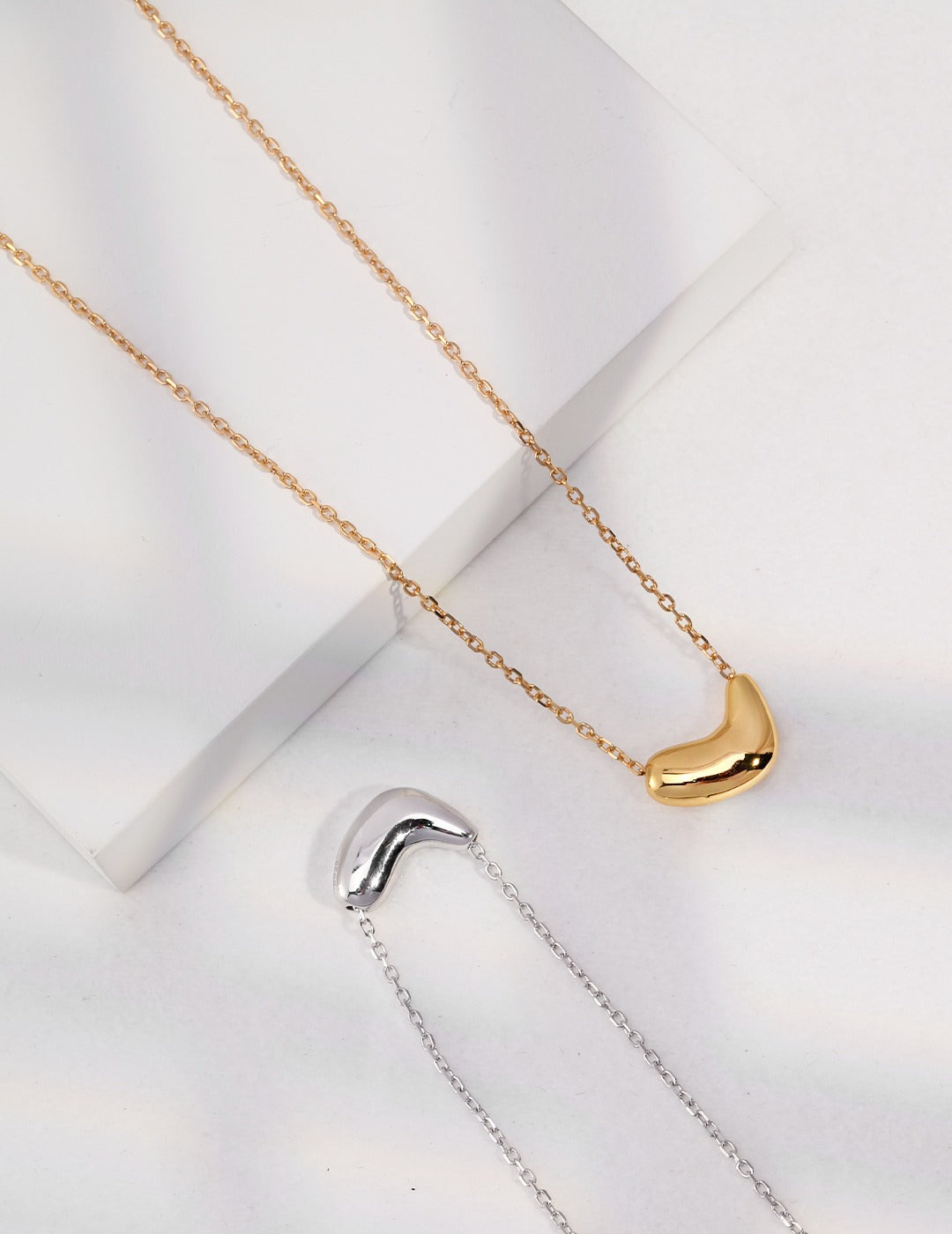 Two bean pendant necklaces in silver and gold with a curved shape.