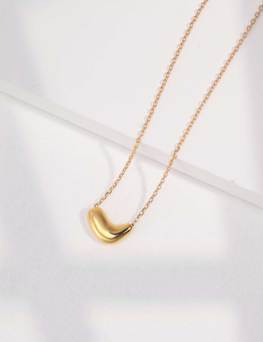 Shiny gold crescent necklace featuring bean pendant on white base.