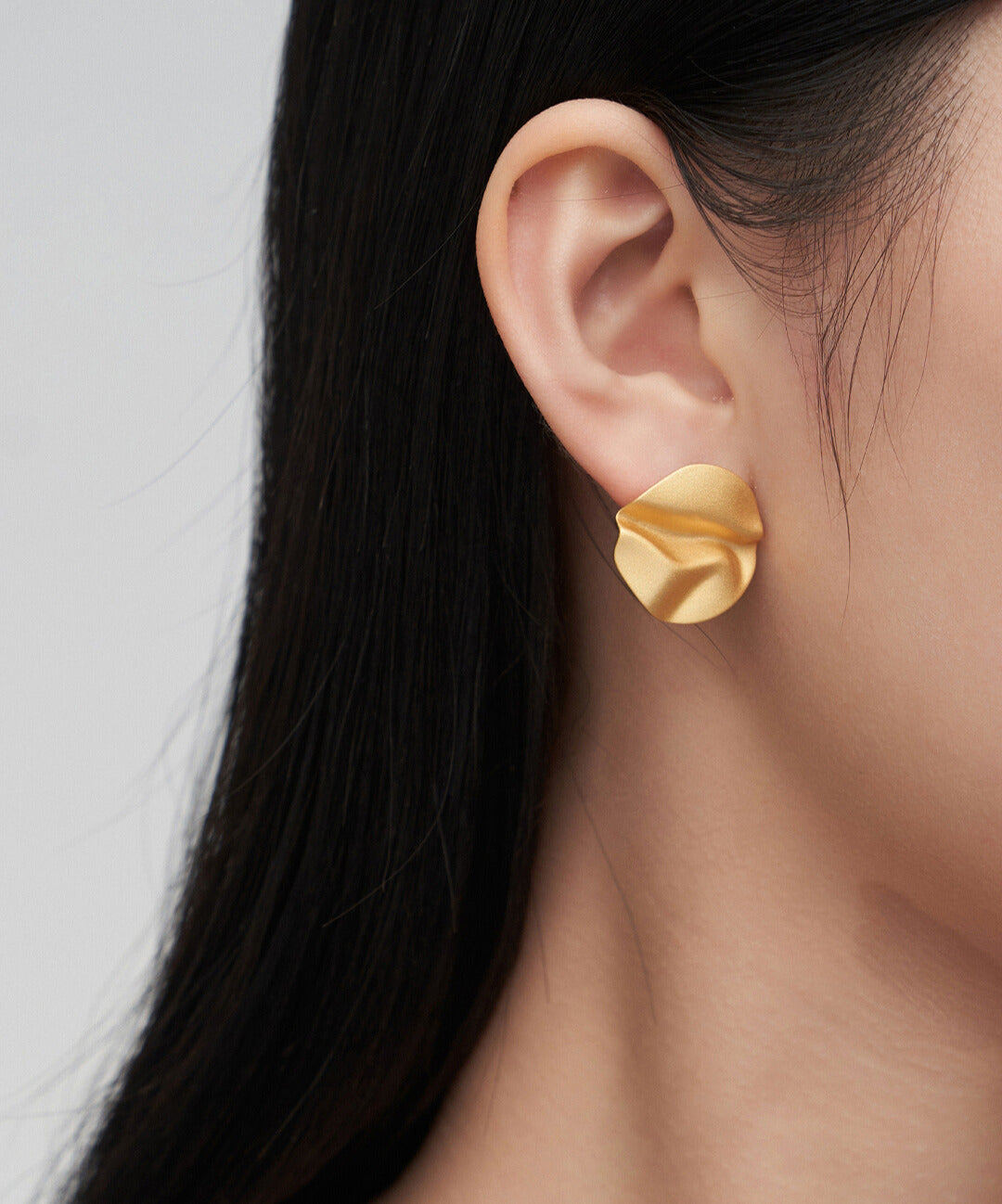 Gold wavy stud earrings shaped like leaves on model's ear.