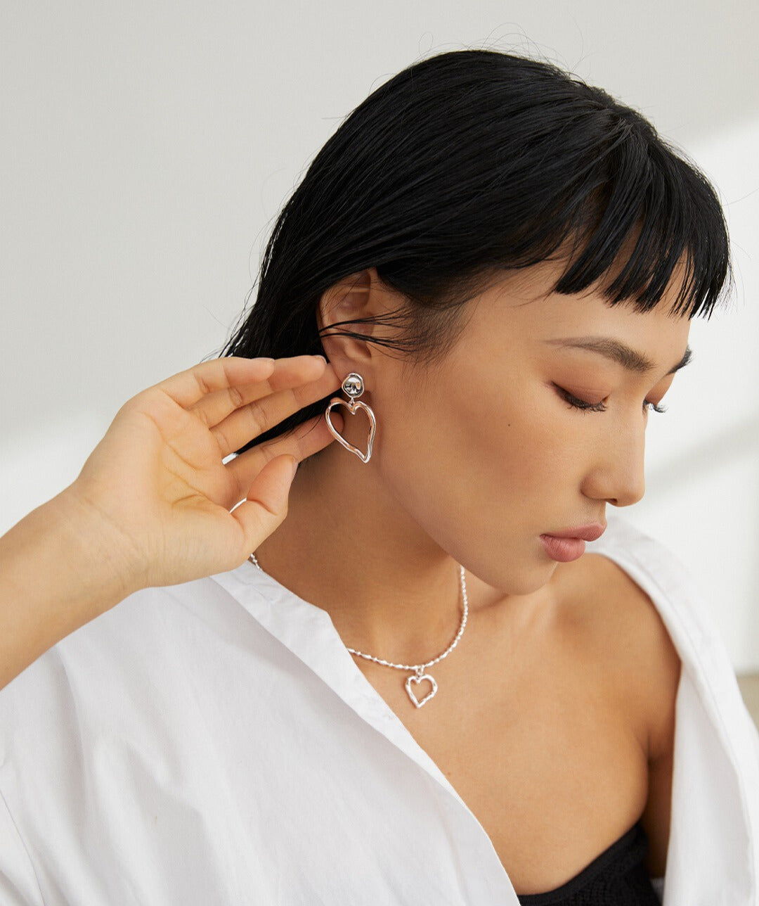 Model wearing silver heart earrings and necklace, adjusting earring.