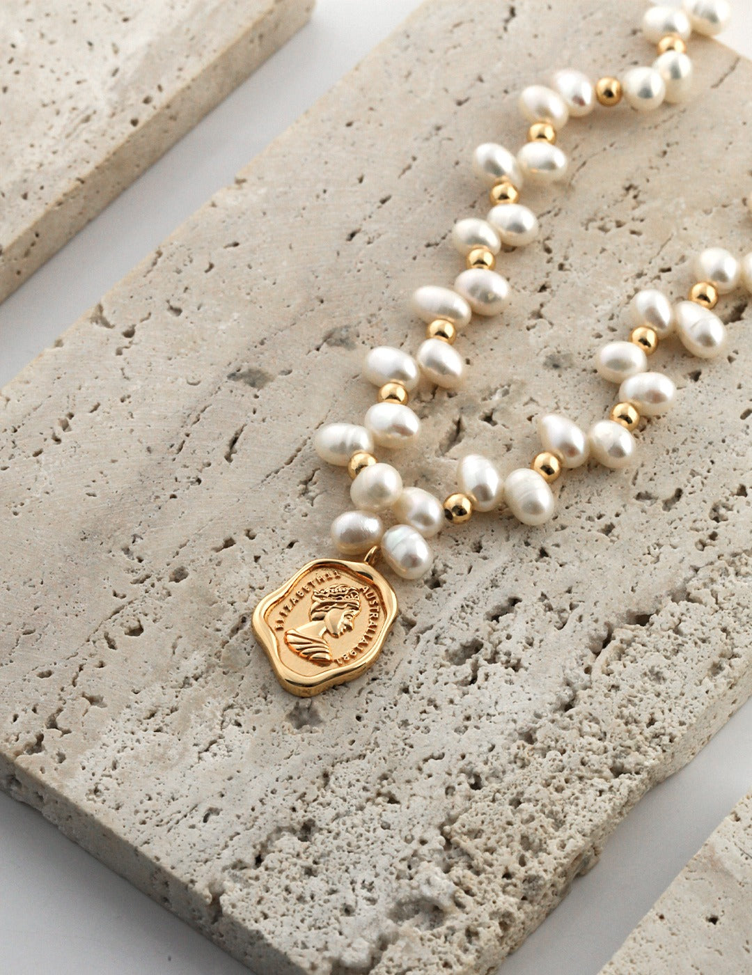 Mezuille Catarina necklace featuring freshwater pearls, gold beads, and a heritage gold coin pendant on a textured stone surface.