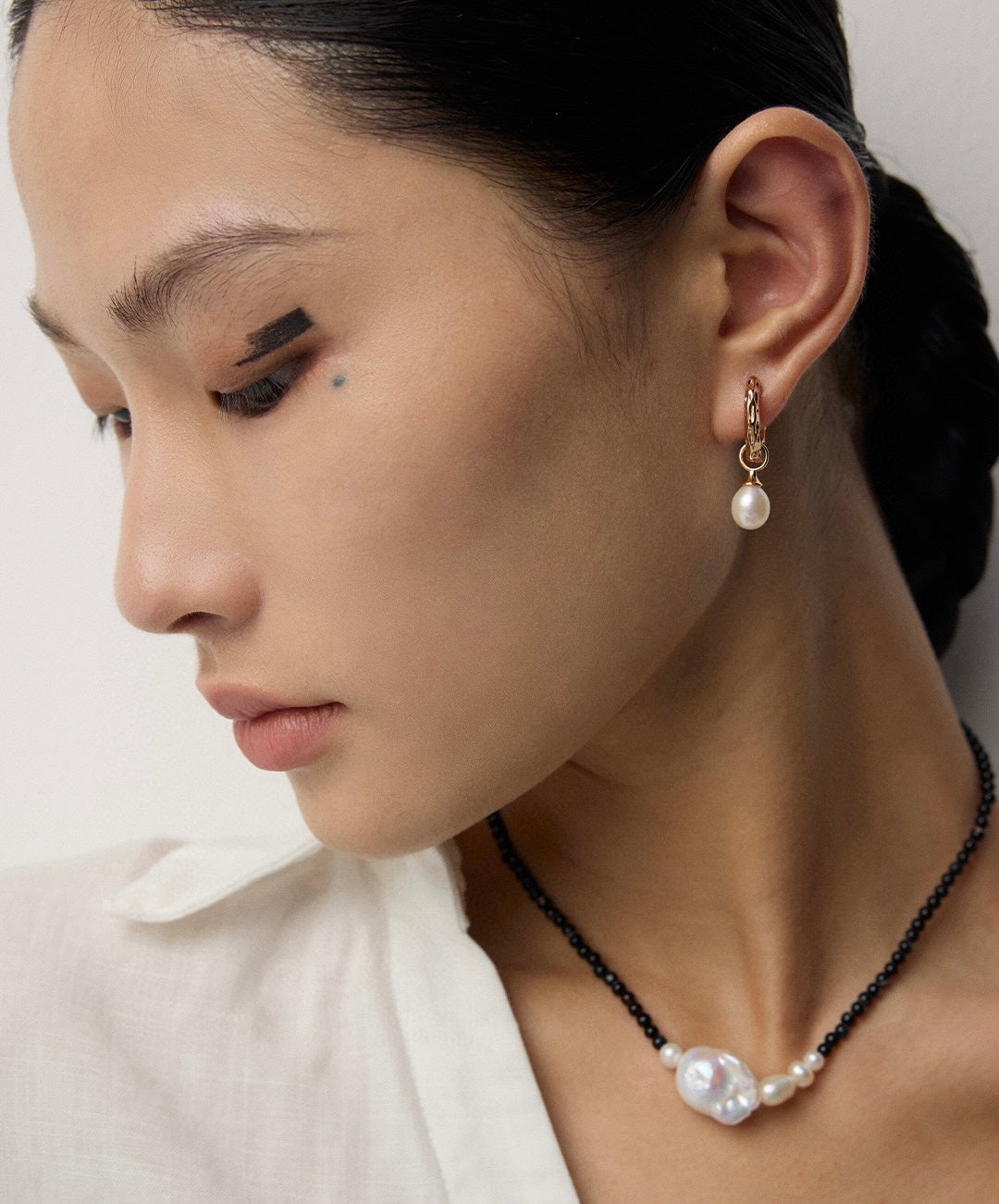 Woman in white shirt wearing minimal gold pearl hoops and a necklace.