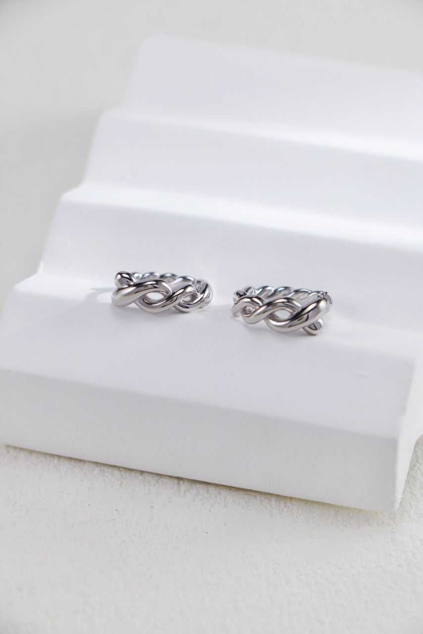 A pair of silver twisted hoop earrings from Mezuille, placed on white surface
