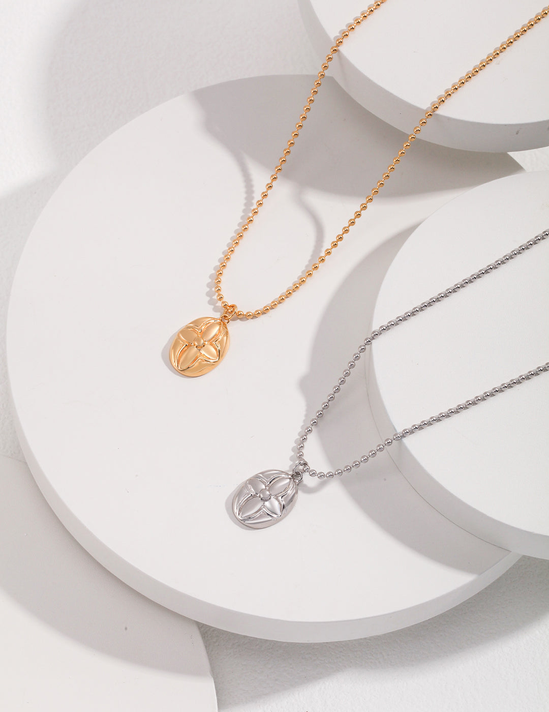 Two necklaces with oval shaped pendants, one gold and one silver, featuring floral patterns engraved on them.